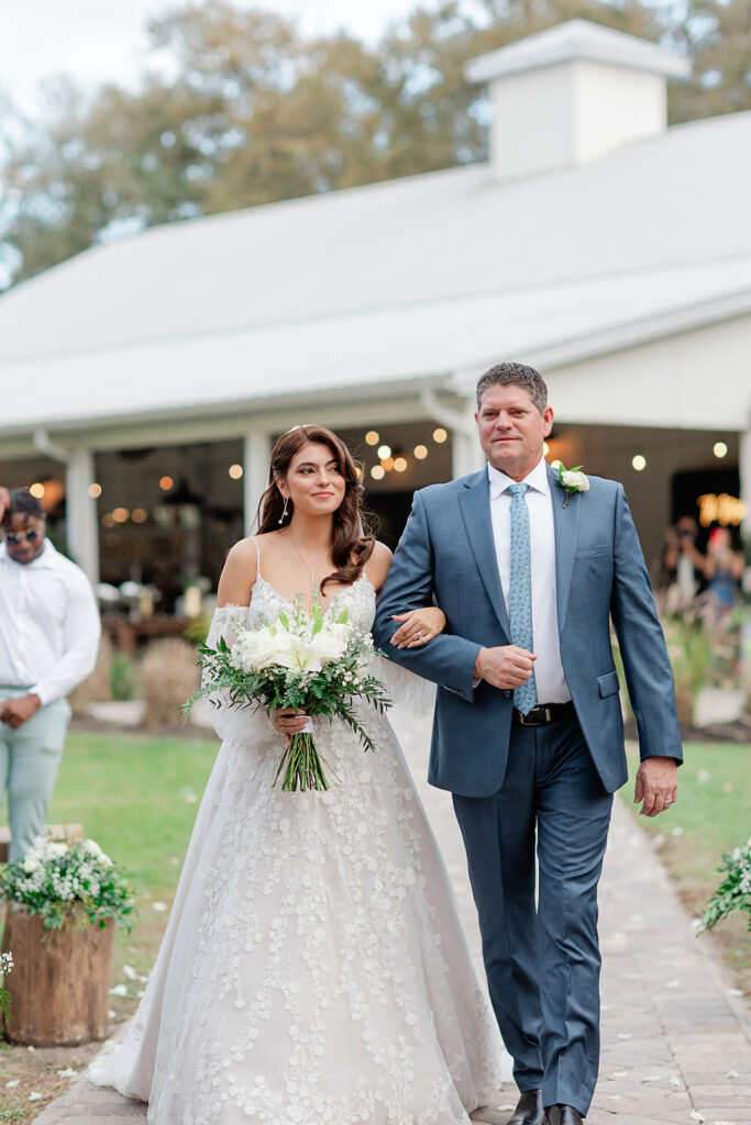 Wedding Photographer in Orlando | Jennifer Holly Light and Airy Wedding Photographer in Orlando | Bride in a white floral gown holding a bouquet walks arm-in-arm with a man in a blue suit. They are outdoors, in front of a rustic venue.
