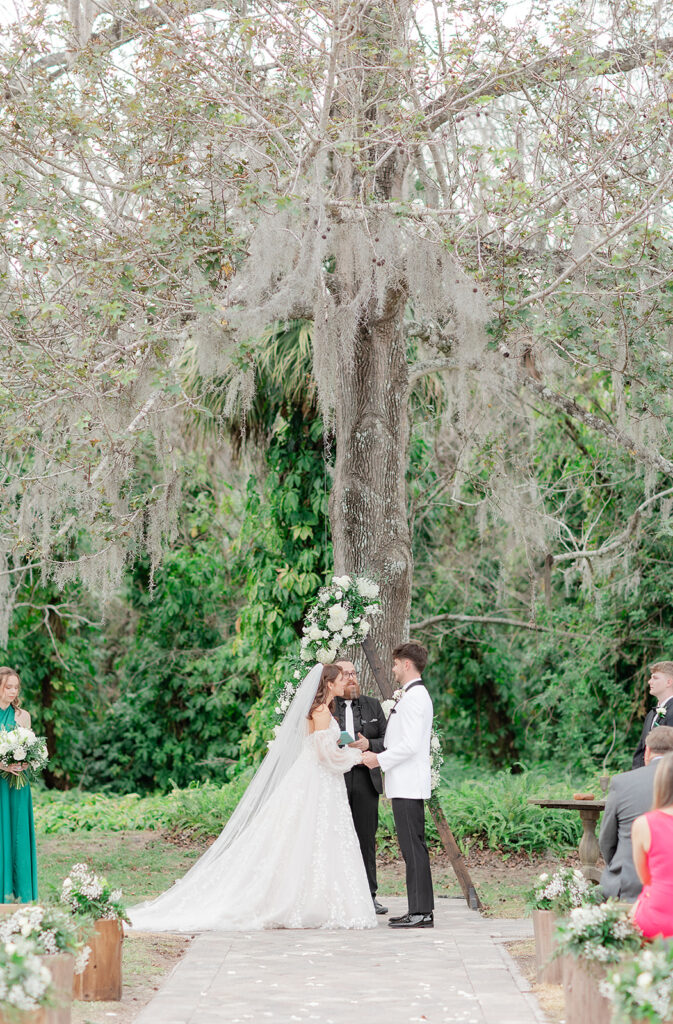 Wedding Photographer in Orlando | Jennifer Holly Light and Airy Wedding Photographer in Orlando | Bride and groom stand under a large tree with floral decorations during an outdoor wedding ceremony. Guests sit on either side, creating a romantic scene.