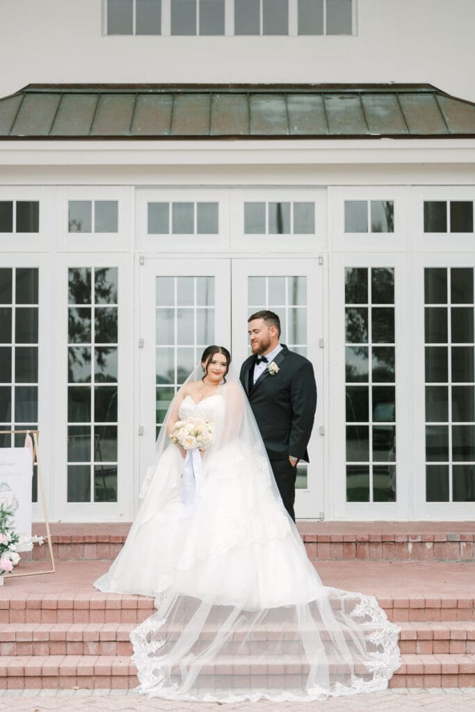 Luxmore Grande Estate | Jennifer Holly Light and Airy Wedding Photographer in Orlando | Bride and groom stand on brick steps in front of elegant doors. Bride in white gown holds bouquet; groom in black suit looks at her affectionately.