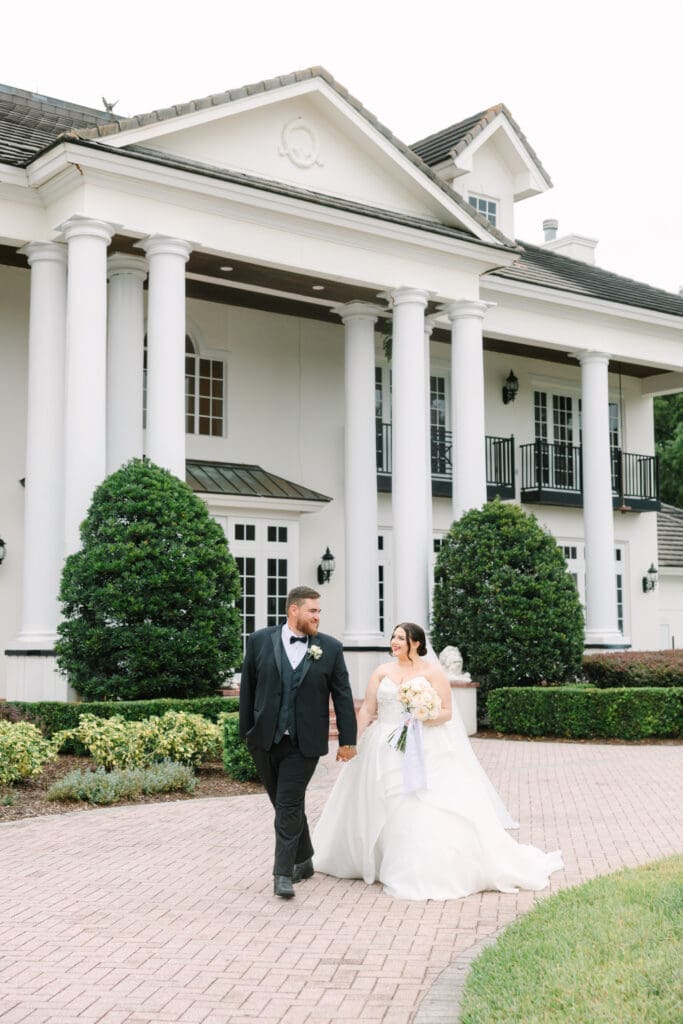 Luxmore Grande Estate | Jennifer Holly Light and Airy Wedding Photographer in Orlando | A newlywed couple walks hand in hand on a cobblestone path in front of an elegant, white-columned mansion. The bride holds a bouquet, and both are smiling.