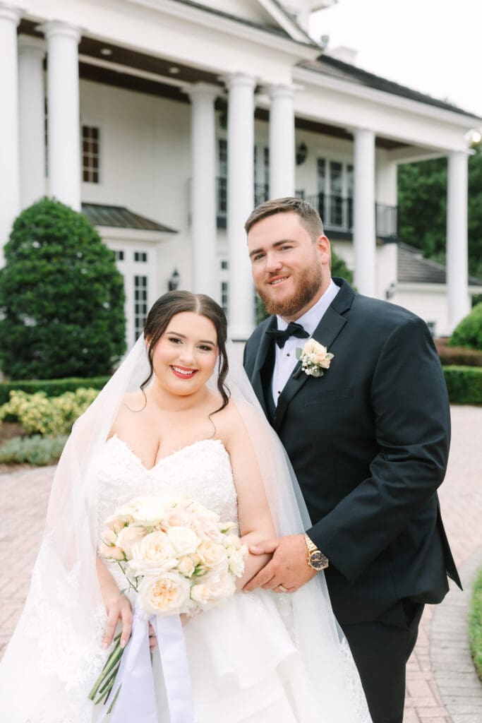 Luxmore Grande Estate | Jennifer Holly Light and Airy Wedding Photographer in Orlando | Bride in white gown holding a bouquet, standing beside a groom in a black tuxedo outside a grand white building. Both are smiling warmly.