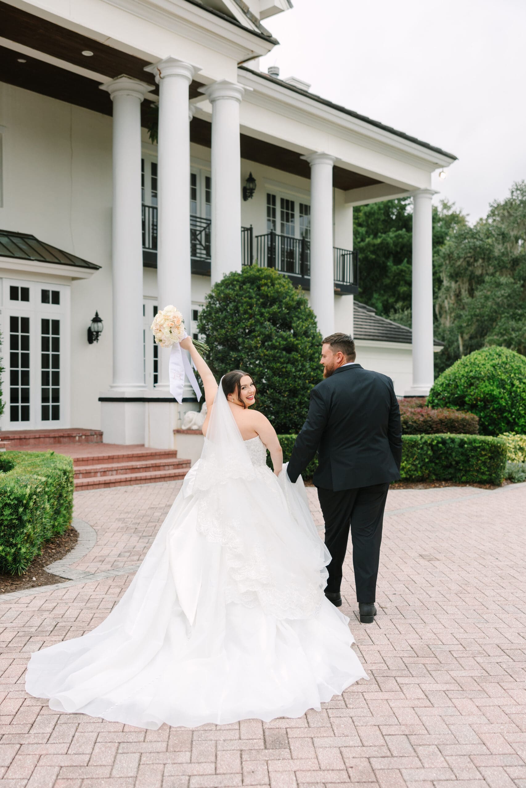 Luxmore Grande Estate | Jennifer Holly Light and Airy Wedding Photographer in Orlando | A bride and groom joyfully walk hand in hand outside a grand white mansion. The bride raises her bouquet, wearing a flowing white gown, exuding happiness.