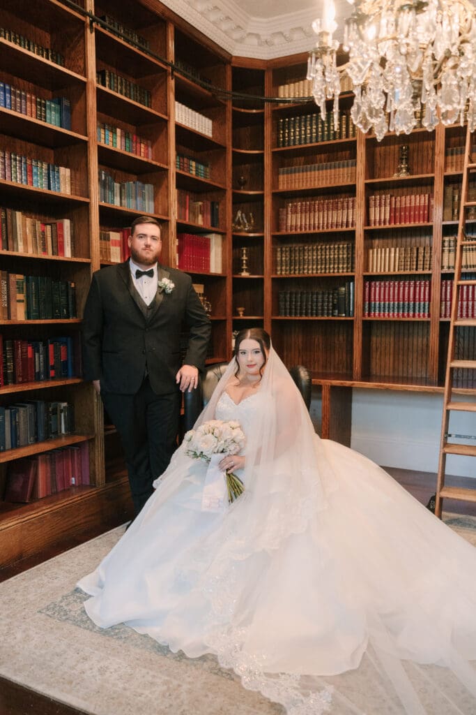 Luxmore Grande Estate | Jennifer Holly Light and Airy Wedding Photographer in Orlando | Bride in elegant white gown seated with bouquet in a classic library. Groom in black tuxedo stands beside her. Ornate chandelier above; warm, timeless ambiance.