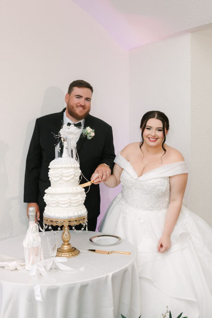 Luxmore Grande Estate | Jennifer Holly Light and Airy Wedding Photographer in Orlando | Bride and groom smile while cutting a tiered white wedding cake. The bride wears an off-shoulder gown; the groom is in a black suit. Joyful moment.
