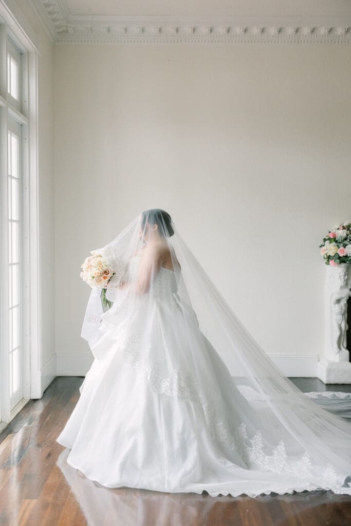 Winter Park Wedding | Jennifer Holly Light and Airy Wedding Photographer in Orlando | Bride in a white wedding gown sits with a long, flowing veil and holds a bouquet of pale flowers, set against a soft, neutral background.