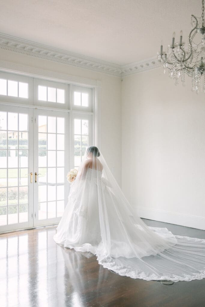 Luxmore Grande Estate | Jennifer Holly Light and Airy Wedding Photographer in Orlando | A bride stands in an elegant, sunlit room with French doors, wearing a flowing white gown and veil, holding a bouquet, under a crystal chandelier.