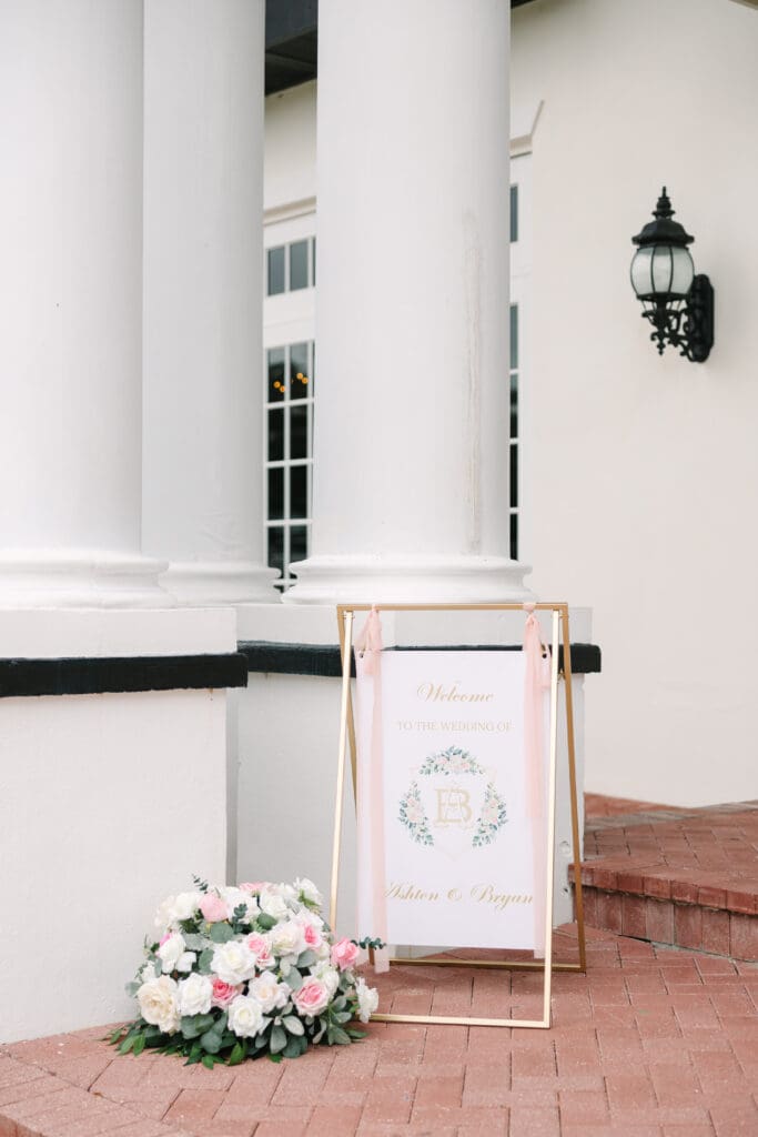 Wedding Photographer in Orlando | Jennifer Holly Light and Airy Wedding Photographer in Orlando | Wedding welcome sign near elegant white columns, framed in gold with soft pink ribbon. A lush bouquet of pink and white roses sits nearby on a brick floor.