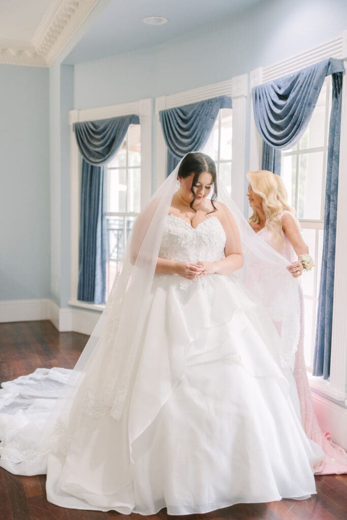 Wedding Photographer Orlando | Jennifer Holly Light and Airy Wedding Photographer in Orlando | A bride in a white gown and veil stands indoors near large windows with blue drapes. A woman assists with the veil, creating a serene, joyful moment.