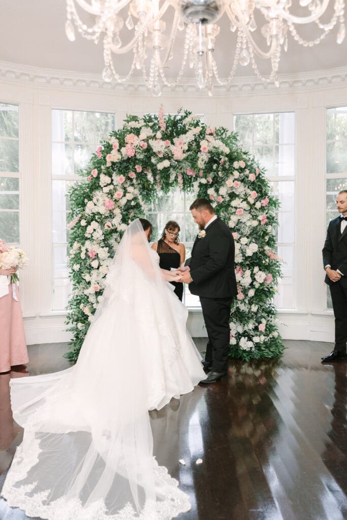 Luxmore Grande Estate | Jennifer Holly Light and Airy Wedding Photographer in Orlando | A couple stands before a lush floral arch, exchanging vows in a bright room with a chandelier. Bride in white gown, groom in black suit, conveying joy and romance.