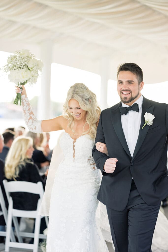 Central Florida Wedding Photographer | Jennifer Holly Light and Airy Wedding Photographer in Orlando | Bride in lace gown joyfully raises bouquet, groom in black tuxedo smiles. They're walking down an aisle with white chairs.