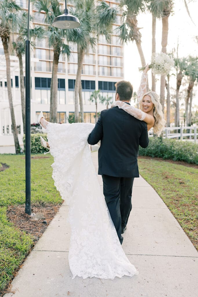 Central Florida Wedding Photographer | Jennifer Holly Light and Airy Wedding Photographer in Orlando | A groom in a black suit carries a smiling bride in a white gown along a garden path with palm trees. The bride holds a bouquet and beams with joy.