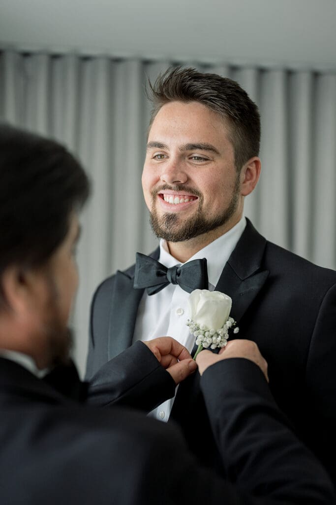 Wedding Photographer Orlando | Jennifer Holly Light and Airy Wedding Photographer in Orlando | Man in a black tuxedo, smiling warmly, has a white boutonniere pinned by another person. The atmosphere is formal and joyful, curtains in the background.