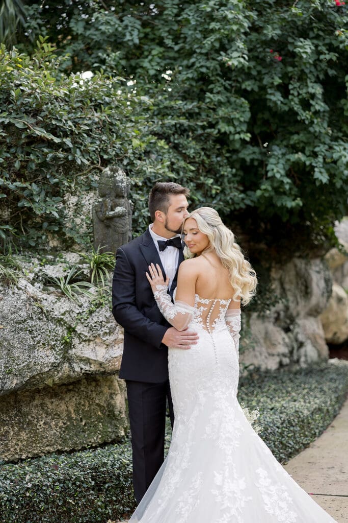 Central Florida Wedding Photographer | Jennifer Holly Light and Airy Wedding Photographer in Orlando | A couple poses outdoors, with the groom in a black suit kissing the bride's forehead. The bride wears a lace-embellished gown, and they stand near lush greenery.