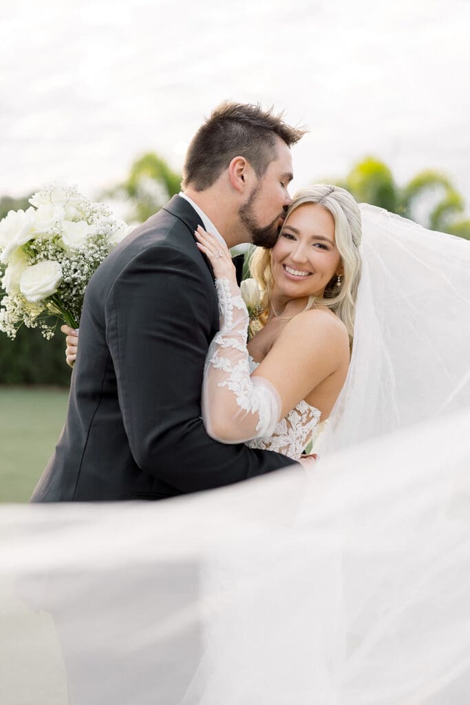 Central Florida Wedding Photographer | Jennifer Holly Light and Airy Wedding Photographer in Orlando | A bride and groom embrace outdoors, smiling warmly. The groom kisses the bride's cheek. She's holding a bouquet of white flowers. Her veil flows in the breeze.