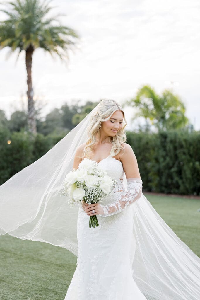 Central Florida Wedding Photographer | Jennifer Holly Light and Airy Wedding Photographer in Orlando | Bride in a lace wedding dress holds a bouquet of white flowers. Her long veil flows behind her. She stands on a lawn with palm trees in the background.
