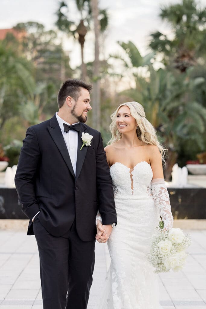 Central Florida Wedding Photographer | Jennifer Holly Light and Airy Wedding Photographer in Orlando | A bride in a lace gown holds white roses, walking hand-in-hand with the groom in a black tuxedo. They smile at each other against a backdrop of palm trees.