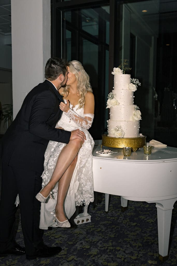 Central Florida Wedding Photographer | Jennifer Holly Light and Airy Wedding Photographer in Orlando | A bride in a white lace dress and a groom in a black suit share a kiss. The bride is sitting on a white grand piano near a tall, decorated wedding cake.