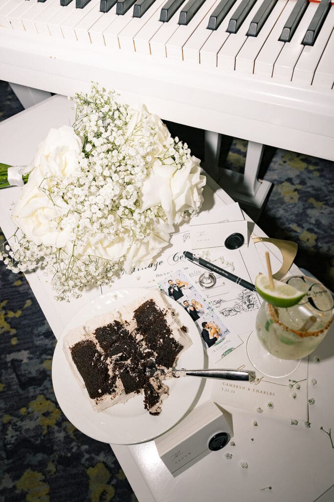 Central Florida Wedding Photographer | Jennifer Holly Light and Airy Wedding Photographer in Orlando | A white piano serves as a backdrop for a wedding scene with a bouquet of white roses and baby's breath, chocolate cake slice, and a cocktail. The mood is elegant and celebratory.