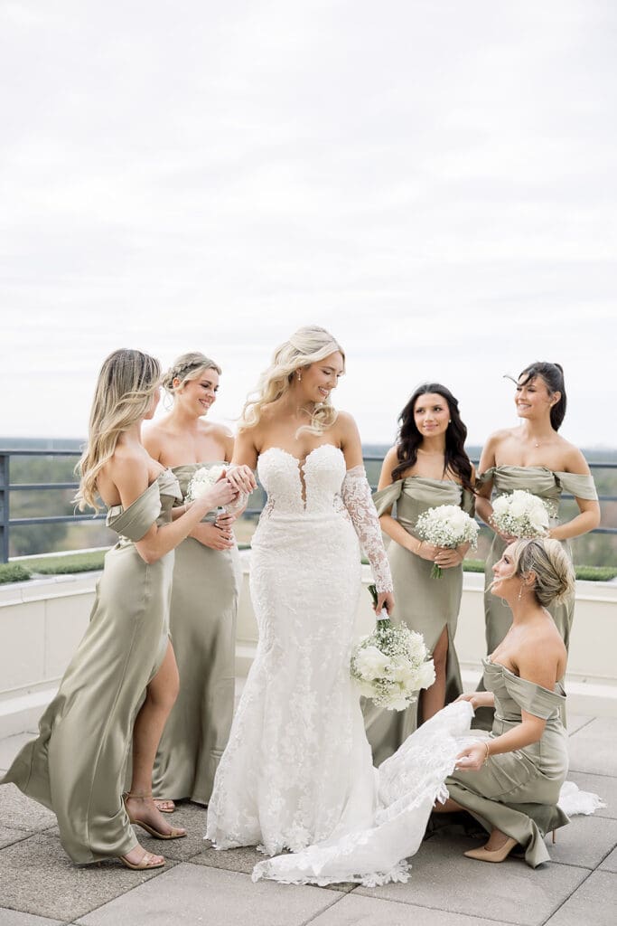 Central Florida Wedding Photographer | Jennifer Holly Light and Airy Wedding Photographer in Orlando | A bride in a lacy white dress stands smiling with five bridesmaids in sage green dresses on a terrace. Everyone is laughing, creating a joyful atmosphere.