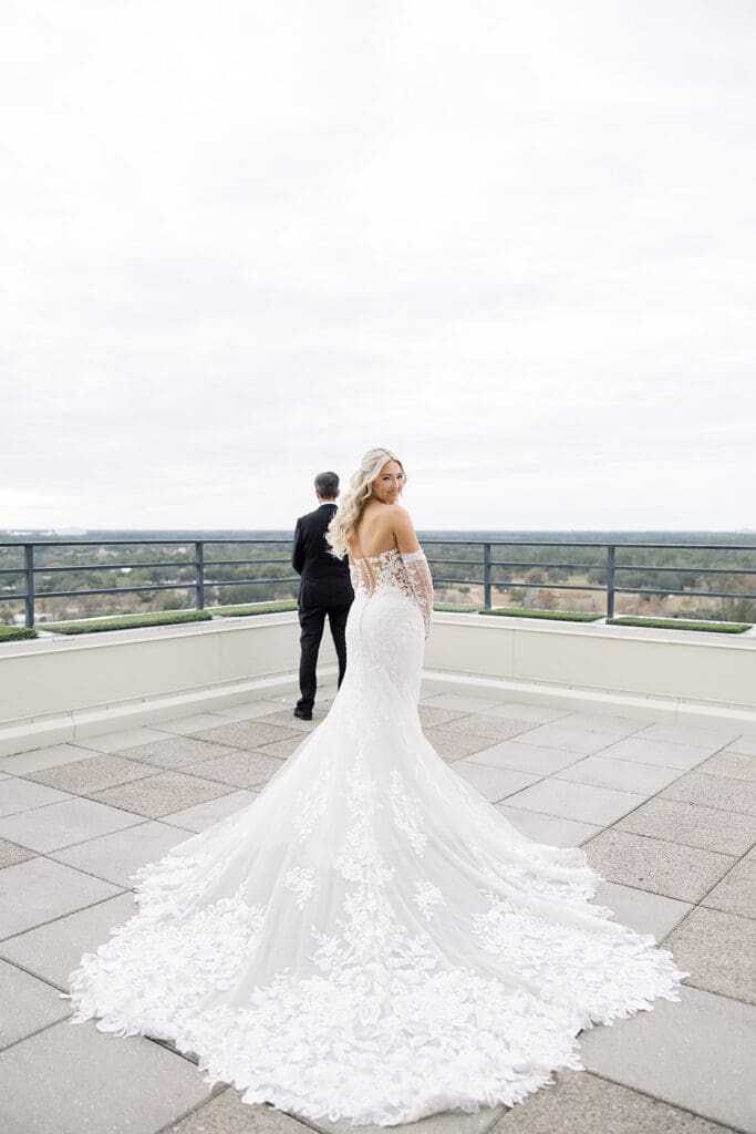 Orlando Wedding Photographer | Jennifer Holly Light and Airy Wedding Photographer in Orlando | A bride in an elegant white gown stands on a rooftop, facing the camera. Her groom, in a black suit, faces away, with a cloudy sky and greenery in the background.