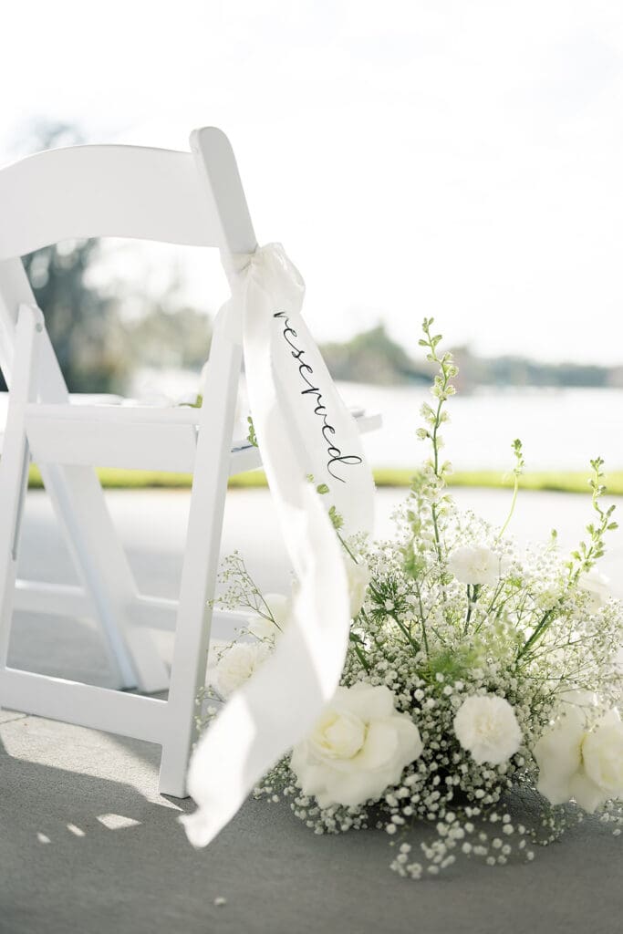 Wedding Photographer Orlando | Jennifer Holly Light and Airy Wedding Photographer in Orlando | White folding chair with a "reserved" ribbon, adorned with delicate white flowers, set against a serene lakeside backdrop. Elegant and tranquil.