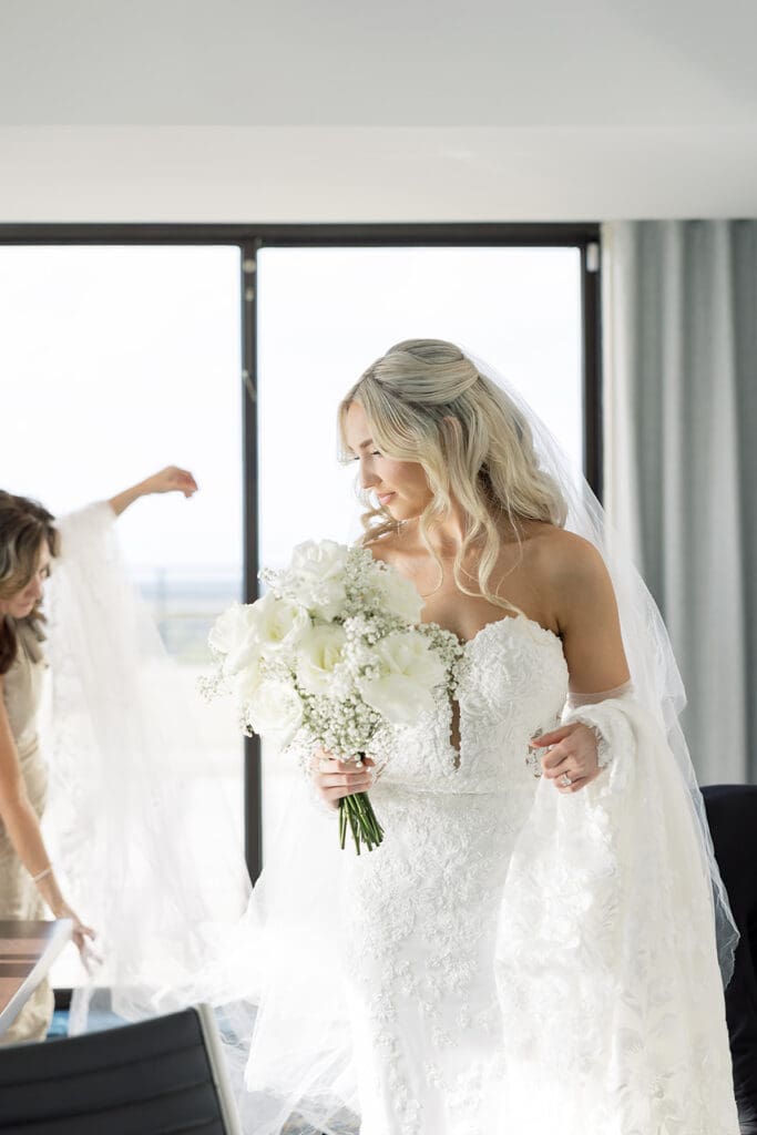 Wedding Photographer Orlando | Jennifer Holly Light and Airy Wedding Photographer in Orlando | Bride in a white lace gown holds a bouquet of white flowers, smiling softly. A bridesmaid adjusts her veil. Bright, airy room with large windows.