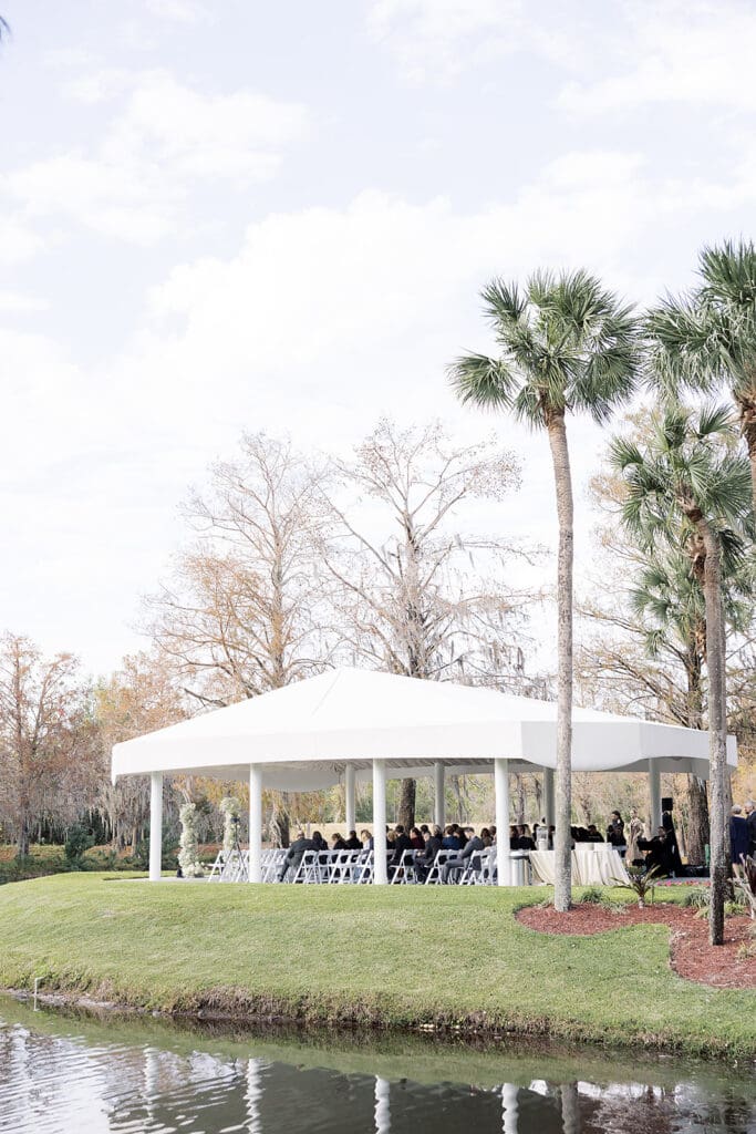 Central Florida Wedding Photographer | Jennifer Holly Light and Airy Wedding Photographer in Orlando | Outdoor event under a white tent on a lush green lawn beside a pond, with guests seated on chairs. Palm trees and bare trees are in the background under a bright sky.