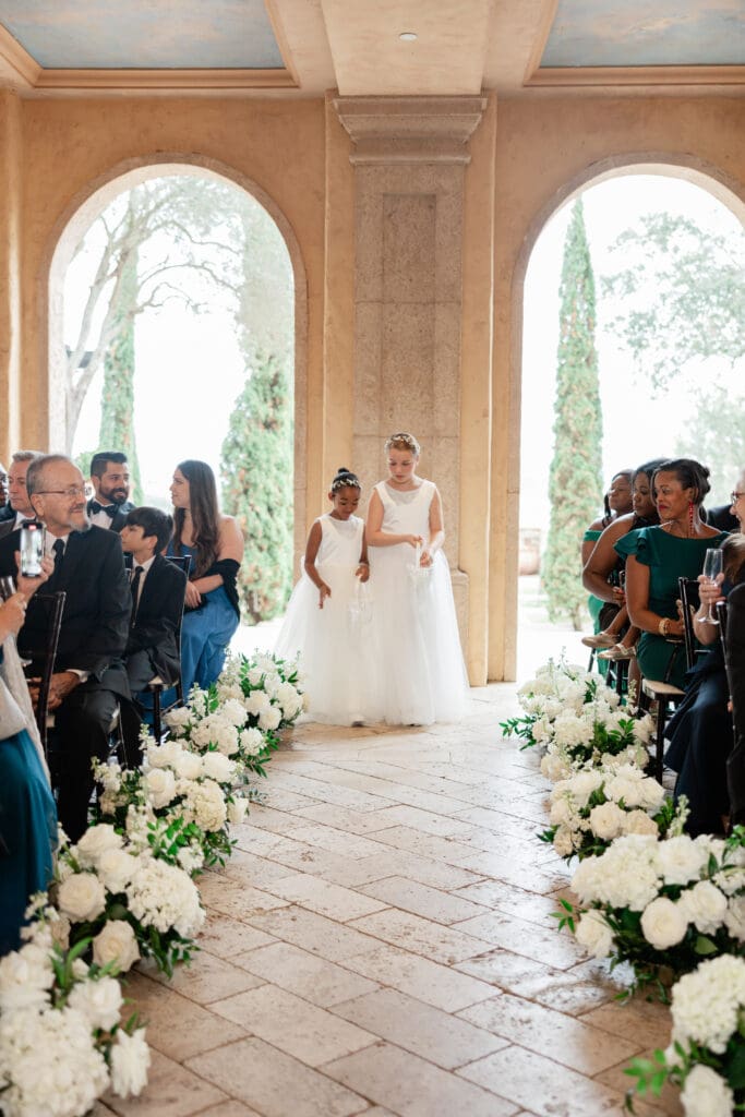 Orlando Wedding Photographer | Jennifer Holly Light and Airy Wedding Photographer in Orlando | Two young girls in white dresses walk down an aisle adorned with white flowers, in a warmly-lit venue. Guests sit on either side watching. Romantic atmosphere.