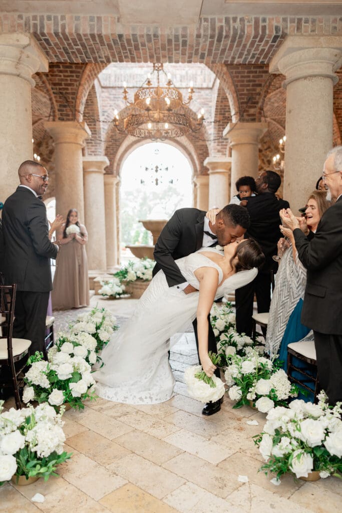 Bella Collina Wedding | Jennifer Holly Light and Airy Wedding Photographer in Orlando | A newlywed couple shares a romantic kiss under a brick archway, surrounded by white floral arrangements. Guests, dressed in formal attire, cheer joyfully.