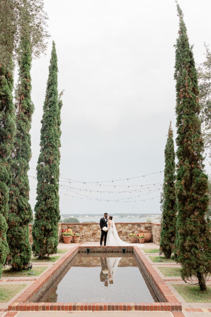 Bella Collina Wedding | Jennifer Holly Light and Airy Wedding Photographer in Orlando | A couple stands at the end of a rectangular reflective pool surrounded by tall cypress trees. String lights hang above, creating a romantic atmosphere.