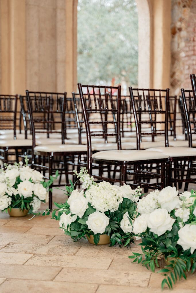 Orlando Wedding Photographer | Jennifer Holly Light and Airy Wedding Photographer in Orlando | Rows of elegant dark wooden chairs with white cushions are arranged for an event. White floral arrangements with greenery line the aisle, exuding a serene and sophisticated ambiance.