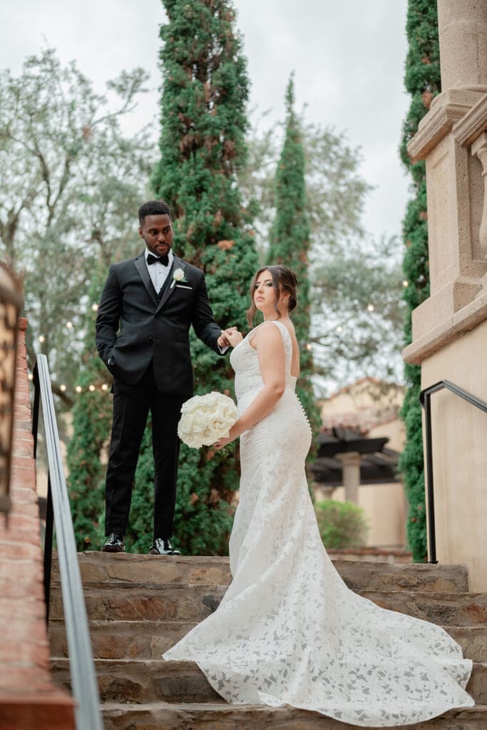 Bella Collina Wedding | Jennifer Holly Light and Airy Wedding Photographer in Orlando | Bride in a lace gown holding flowers stands on stone steps, looking upward. Groom in a black suit stands nearby, set against lush greenery.