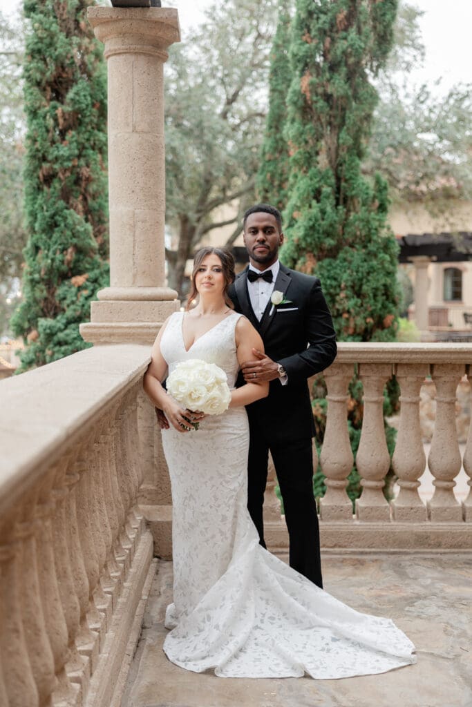 Bella Collina Wedding | Jennifer Holly Light and Airy Wedding Photographer in Orlando | A bride in a lace gown holds white roses, while the groom in a black tuxedo stands behind her on a stone balcony, with green trees in the background.