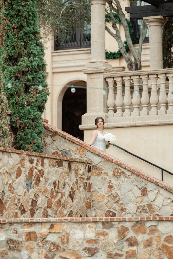 Wedding Photographer in Orlando | Jennifer Holly Light and Airy Wedding Photographer in Orlando | A bride in a white gown holds a bouquet, standing on a stone staircase beside a lush green tree, with elegant balustrades in the background.