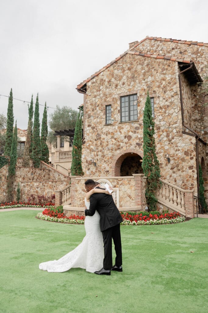 Bella Collina Wedding | Jennifer Holly Light and Airy Wedding Photographer in Orlando | Bride and groom embrace in front of a rustic stone building with a lush green lawn and vibrant flower bed.
