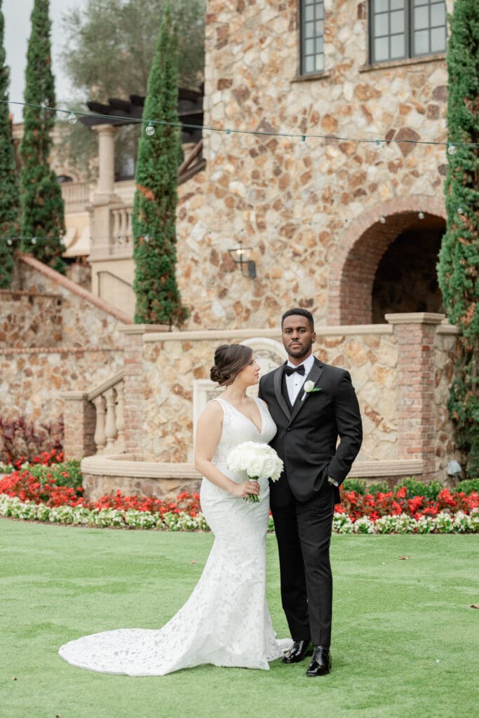 Orlando Wedding Photographer | Jennifer Holly Light and Airy Wedding Photographer in Orlando | Bride in lace gown holds white bouquet, standing with groom in black tuxedo. They are in front of a stone building with greenery and flowers around.