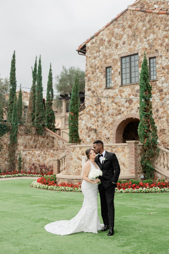 Orlando Wedding Photographer | Jennifer Holly Light and Airy Wedding Photographer in Orlando | A bride and groom share a kiss in front of a stone building with lush, green cypress trees and colorful flower beds, emanating love and joy.