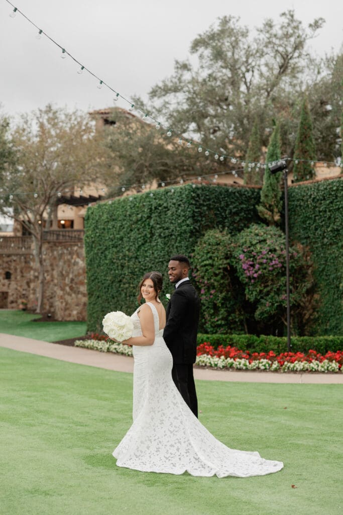 Bella Collina Wedding | Jennifer Holly Light and Airy Wedding Photographer in Orlando | A bride in a white lace gown holds a bouquet, smiling at the camera alongside a groom in a black suit. They're outdoors on lush grass, near a groomed hedge.
