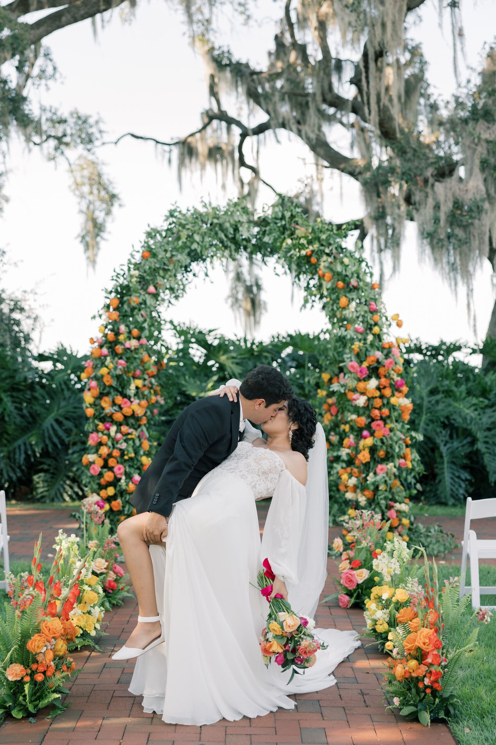 Garden Wedding Venues in Orlando | Jennifer Holly Light and Airy Wedding Photographer in Orlando | A couple shares a romantic kiss under a flower arch, amidst vibrant orange and pink blooms. The bride holds a bouquet, exuding joy and celebration.