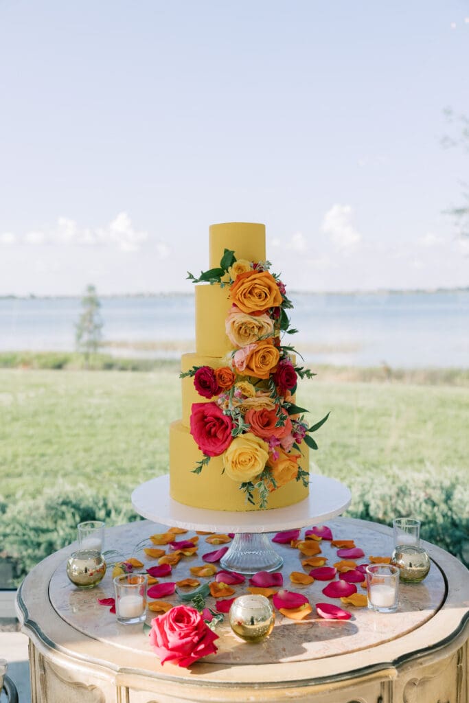Garden Wedding Venues in Orlando | Jennifer Holly Light and Airy Wedding Photographer in Orlando | A three-tiered yellow cake adorned with vibrant red and orange roses stands on a table scattered with flower petals. A serene lake and green grass create a picturesque backdrop.