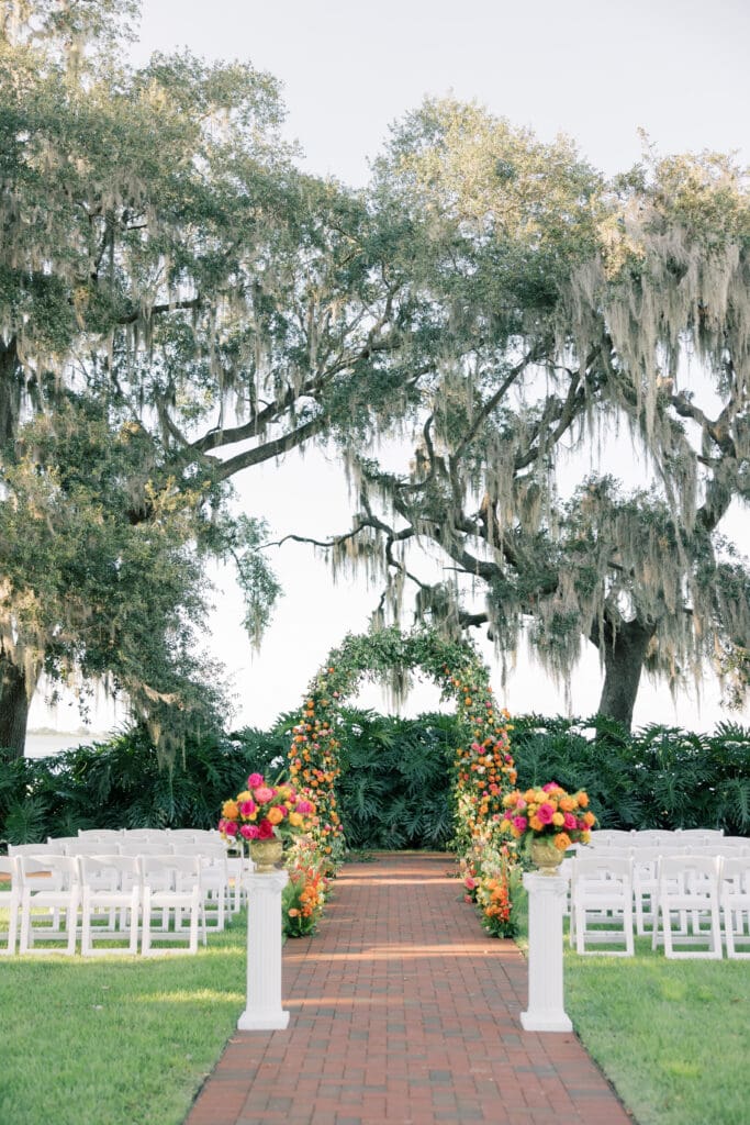 Garden Wedding Venues in Orlando | Jennifer Holly Light and Airy Wedding Photographer in Orlando | Wedding ceremony setting with a floral archway on a red-brick path, flanked by white chairs and flower arrangements. Towering oak trees frame the scene.