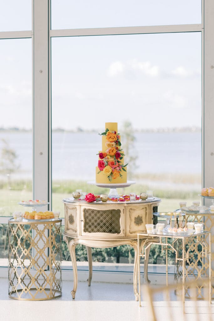 Winter Park Wedding | Jennifer Holly Light and Airy Wedding Photographer in Orlando | A three-tiered yellow cake adorned with bright flowers sits on an ornate table by a large window with a serene lake view. Elegant and vibrant setting.