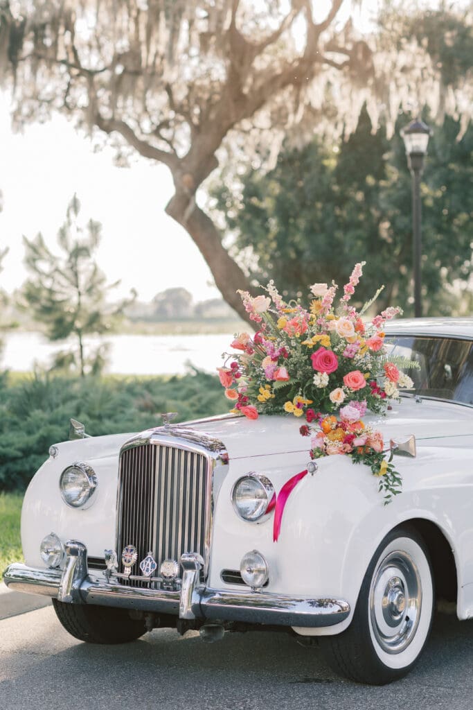 Garden Wedding Venues in Orlando | Jennifer Holly Light and Airy Wedding Photographer in Orlando | Vintage white car adorned with a vibrant floral arrangement of pink, yellow, and orange flowers on its front. Background includes trees and soft sunlight. Romantic and elegant atmosphere.