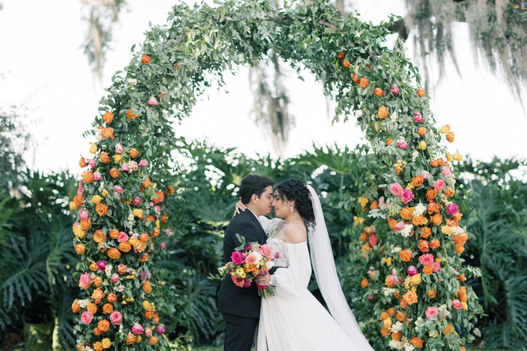 Garden Wedding Venues in Orlando | Jennifer Holly Light and Airy Wedding Photographer in Orlando | A couple embraces under a lush, leafy arch adorned with vibrant orange and pink flowers. The bride holds a colorful bouquet, conveying a romantic, serene atmosphere.