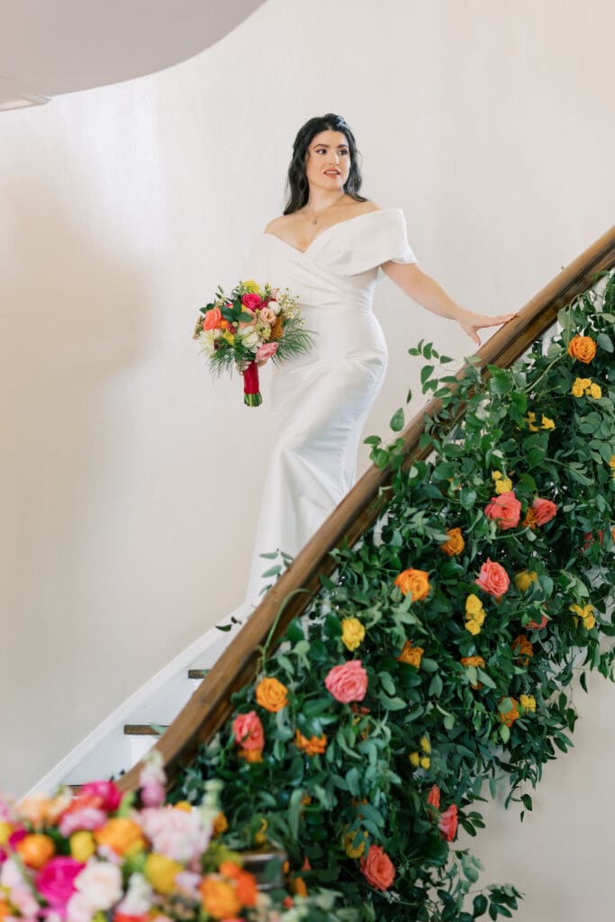 Garden Wedding Venues in Orlando | Jennifer Holly Light and Airy Wedding Photographer in Orlando | Bride in an elegant white gown descends a floral-adorned staircase, holding a vibrant bouquet. The scene exudes joy and elegance against a soft, neutral background.
