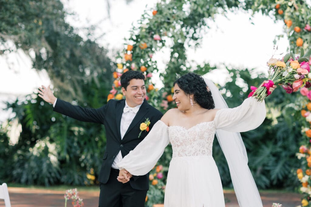 Garden Wedding Venues in Orlando | Jennifer Holly Light and Airy Wedding Photographer in Orlando | A joyful couple celebrates outdoors against a floral arch backdrop. The bride wears a lace dress and veil, and the groom is in a black suit.