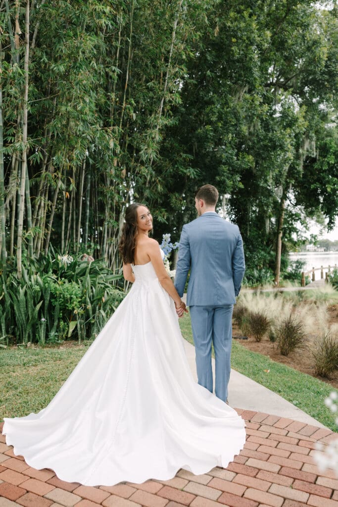 Outdoor Wedding Venues Orlando | Jennifer Holly Light and Airy Wedding Photographer in Orlando | A bride in a flowing white gown and a groom in a blue suit hold hands, walking on a brick path surrounded by lush greenery and trees. The scene is serene and romantic.