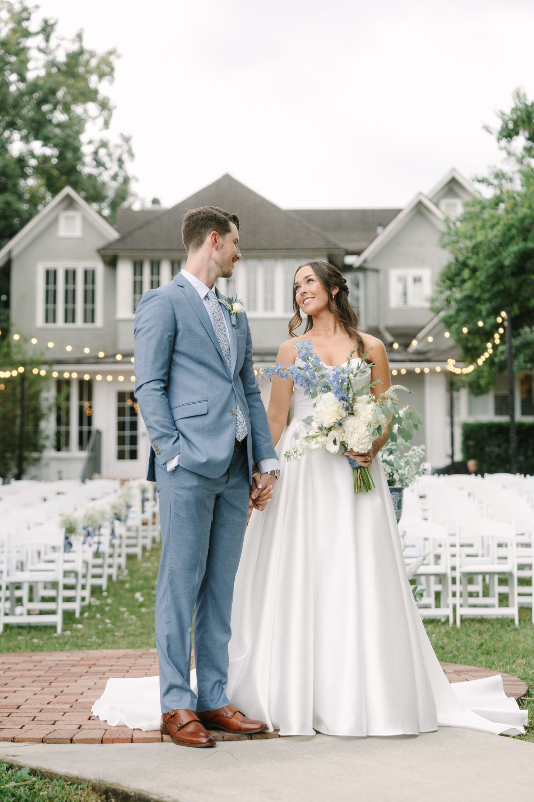 Wedding Venues Winter Park | Jennifer Holly Light and Airy Wedding Photographer in Orlando | A couple stands hand in hand, gazing at each other, in front of a charming house with fairy lights. The bride holds a bouquet of white and blue flowers.