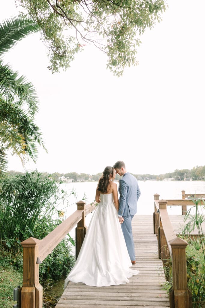 Winter Park Wedding | Jennifer Holly Light and Airy Wedding Photographer in Orlando | A couple stands on a wooden pier holding hands, facing a serene lake. The bride wears a flowing white gown, while the groom is in a light blue suit. Lush greenery surrounds them, conveying a romantic and peaceful atmosphere.