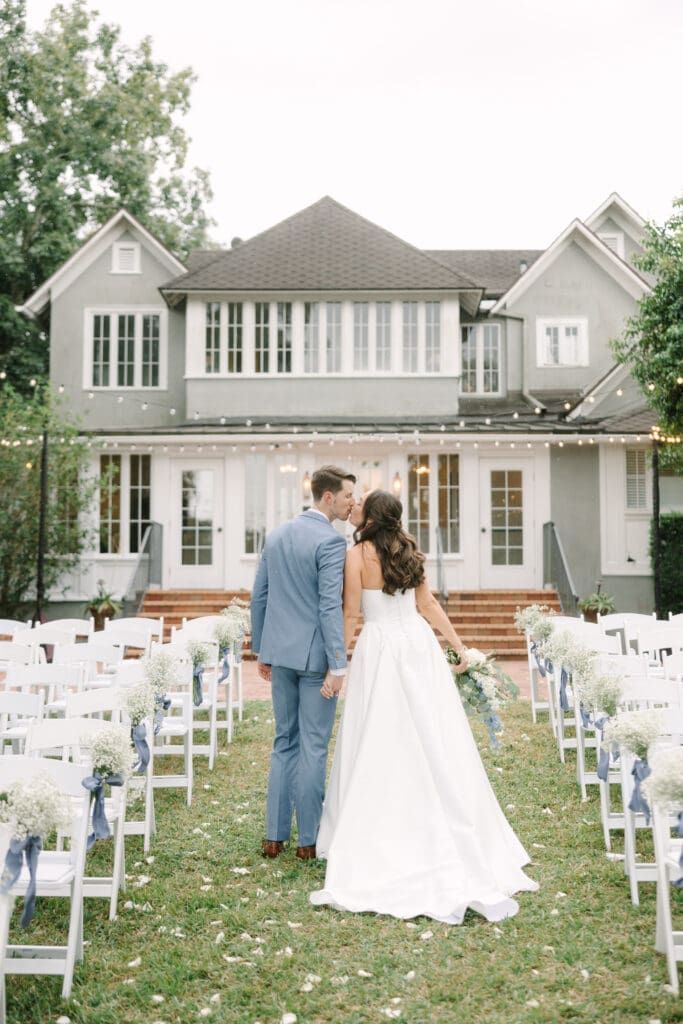 Outdoor Wedding Venues Orlando | Jennifer Holly Light and Airy Wedding Photographer in Orlando | Bride and groom walk down a grassy aisle, holding hands, towards a large white house adorned with string lights. The scene exudes romance and elegance.